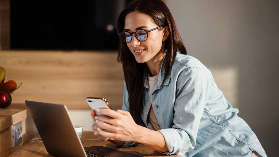 Een vrouw met een bril kijkt naar een smartphone bij een laptop op een houten tafel.