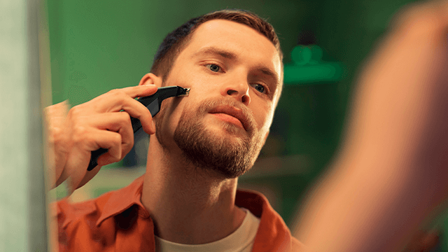 Een man met een oranje shirt trimt zijn baard met een zwart apparaat voor een groene achtergrond.