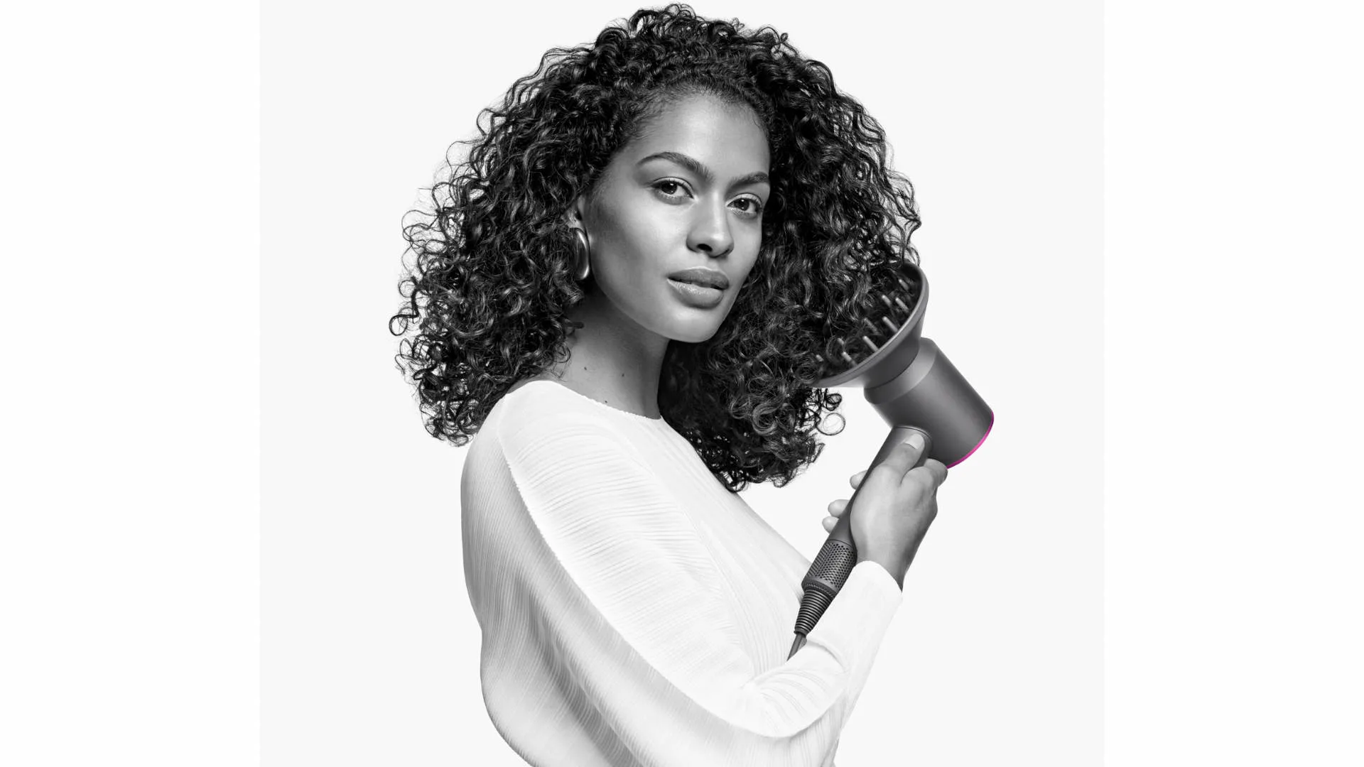 Een vrouw met krullend haar houdt een grijze föhn met een diffuser vast tegen een witte achtergrond.