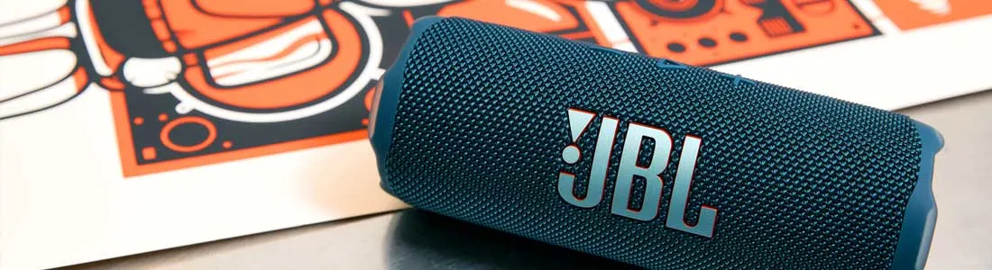 Blauwe JBL-bluetoothspeaker ligt schuin op een tafel met een kleurrijke poster op de achtergrond.