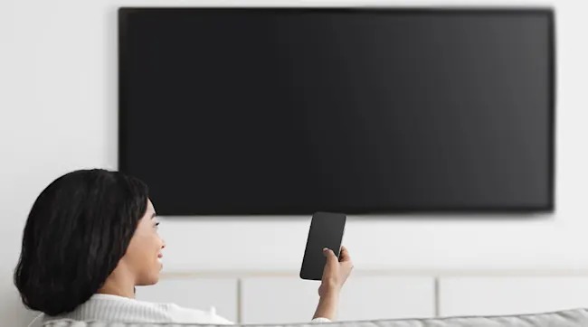 Een vrouw zit op een bank voor een grote zwarte tv aan een witte muur en houdt een zwarte afstandsbediening vast.