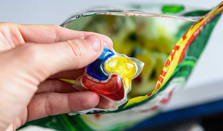A hand holds a colourful dishwasher tablet above an opened bag, ready for use in the dishwasher.