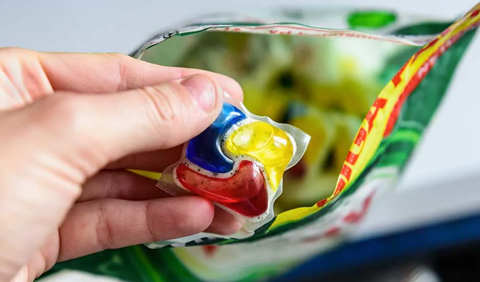 Een hand houdt een kleurrijke vaatwastablet vast boven een geopende zak, klaar voor gebruik in de afwasmachine.