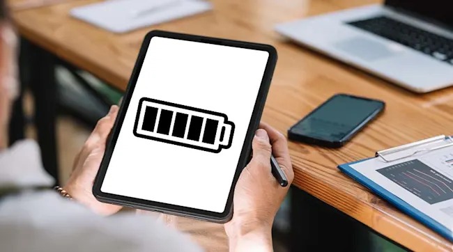 Persoon houdt een tablet vast met een batterijsymbool. Op de houten tafel ligt een telefoon, laptop en klembord.