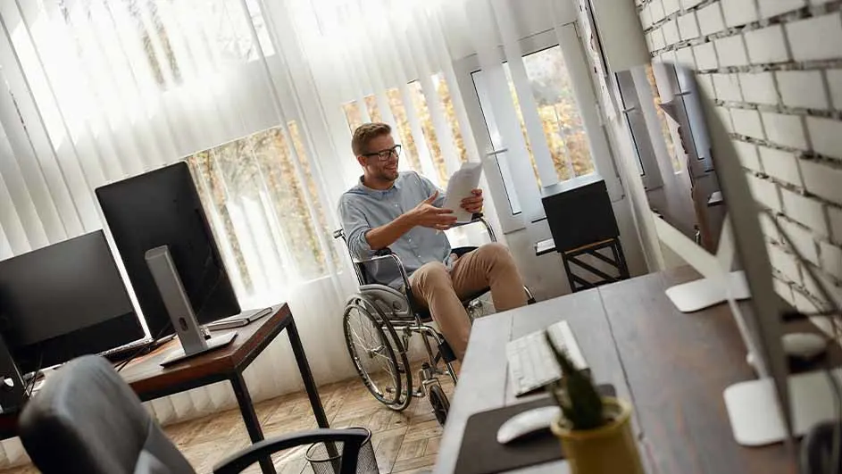 Een man in een rolstoel bekijkt documenten in een kantoor met bureaus en computers.