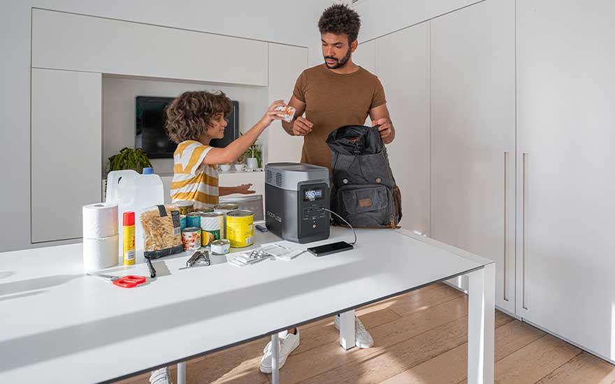 Vader en kind vullen samen een rugzak met proviand aan een tafel waar een portable stroomgenerator en noodartikelen klaarliggen, in een heldere kamer.