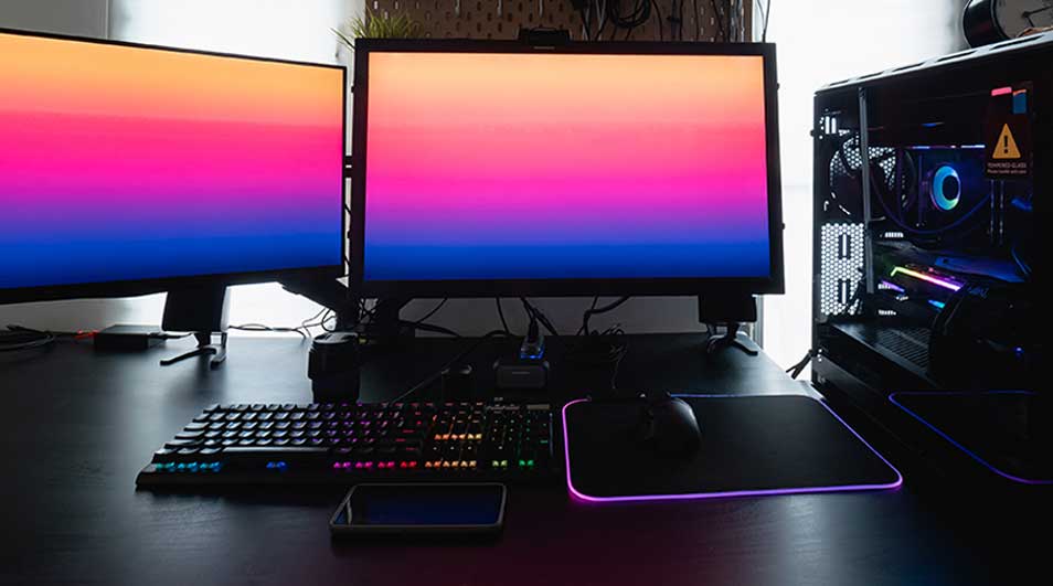 Strakke gamingopstelling met drie schermen, RGB-toetsenbord, muismat en verlichte pc-kast op een donker bureau.