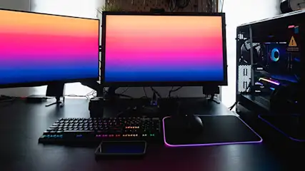 Strakke gamingopstelling met drie schermen, RGB-toetsenbord, muismat en verlichte pc-kast op een donker bureau.