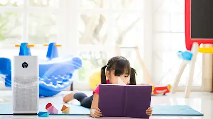 Meisje leest paars boek liggend op een blauwe mat. Speelgoed, een wit apparaat, en een krijtbord zijn zichtbaar.