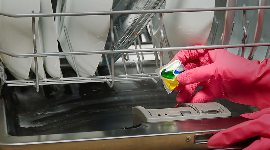 Handen met roze handschoenen plaatsen een vaatwastablet in de vaatwasser, met witte borden in het rek.
