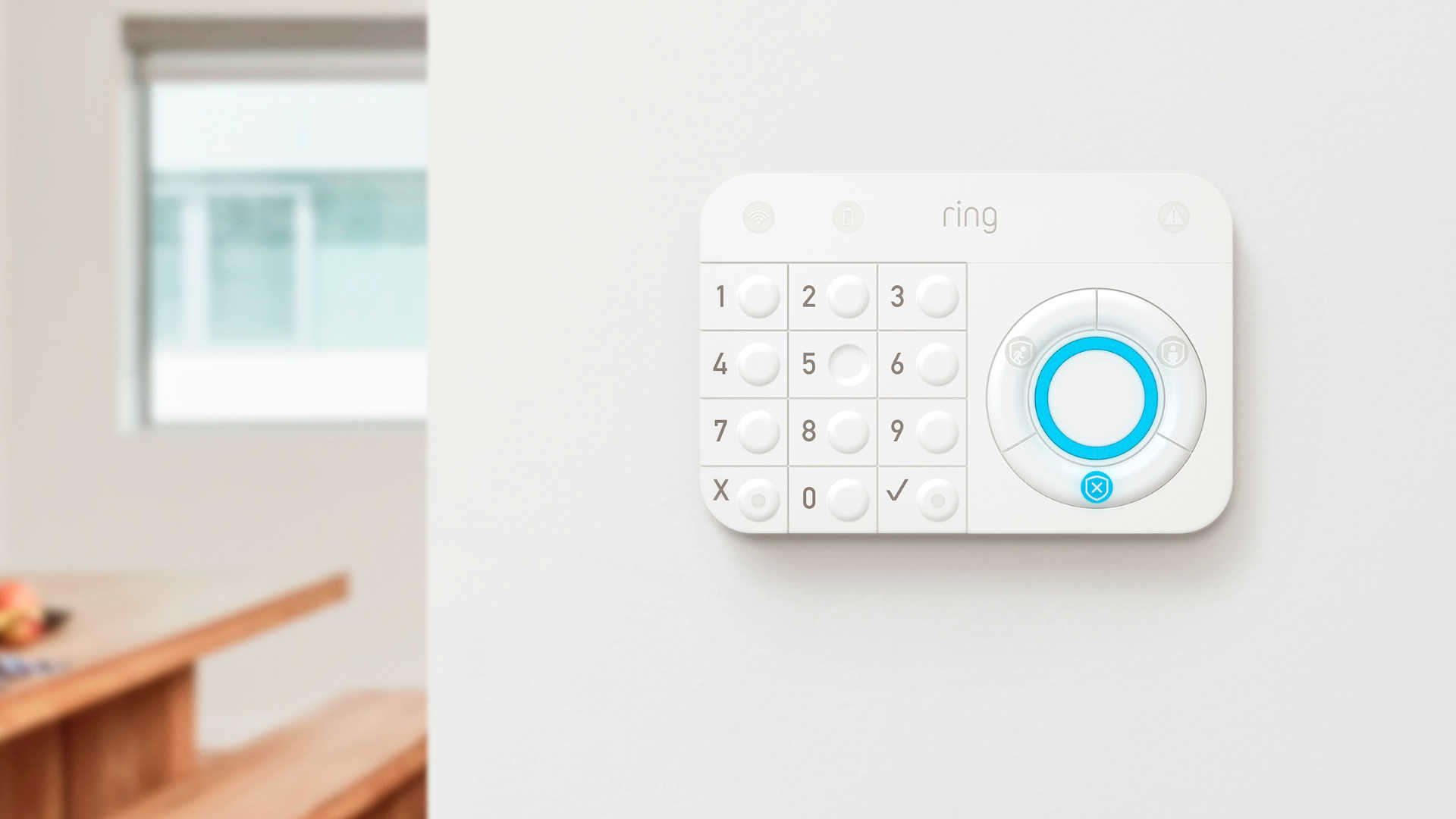 Een wit Ring-alarmtoetsenbord hangt aan een witte muur, met een raam en een houten tafel op de achtergrond.