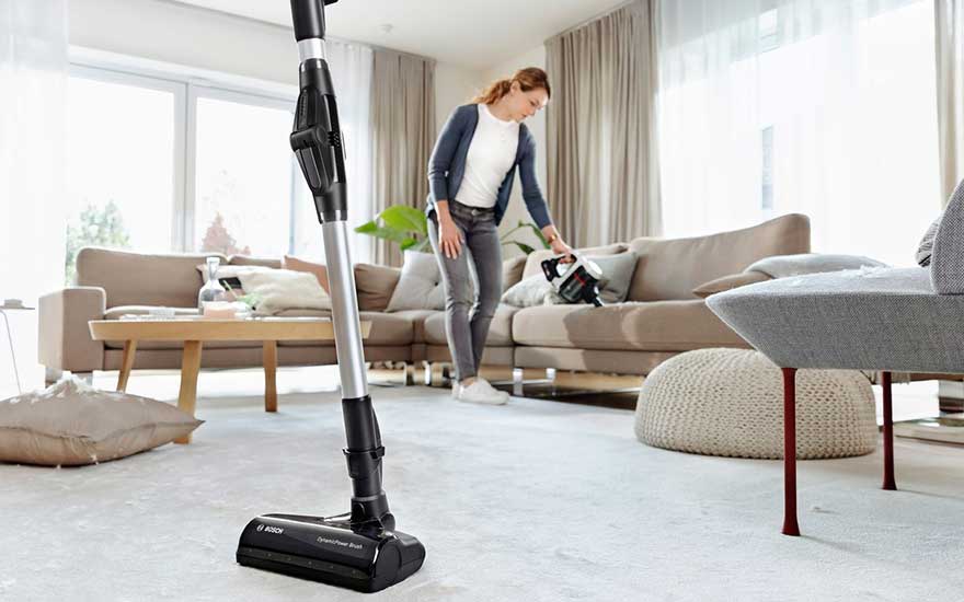 Een vrouw gebruikt een steelstofzuiger in haar woonkamer om het vloerkleed schoon te maken, met een lichte en moderne uitstraling.