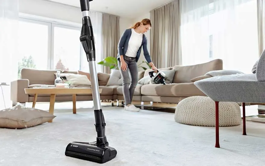 Een vrouw gebruikt een steelstofzuiger in haar woonkamer om het vloerkleed schoon te maken, met een lichte en moderne uitstraling.