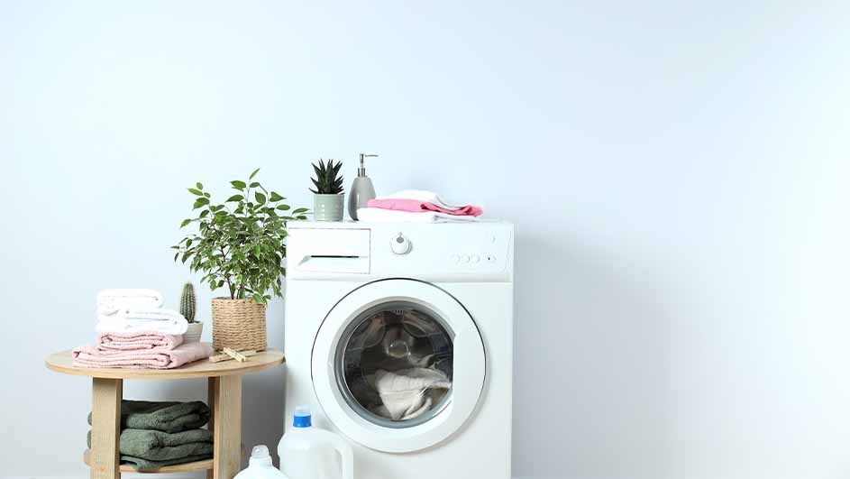 A white washing machine with a round door is surrounded by folded towels, green plants, and detergent, creating a fresh laundry room look.