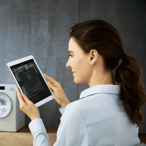 Vrouw bekijkt een tablet in een kamer met een wasmachine op de achtergrond. Ze draagt een wit overhemd en haar haar in een staart.