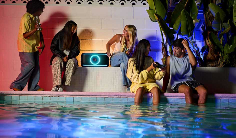 Vijf jongeren feesten bij een zwembad, twee met voeten in het water, kleurrijke lichten en een grote luidspreker tussen hen.