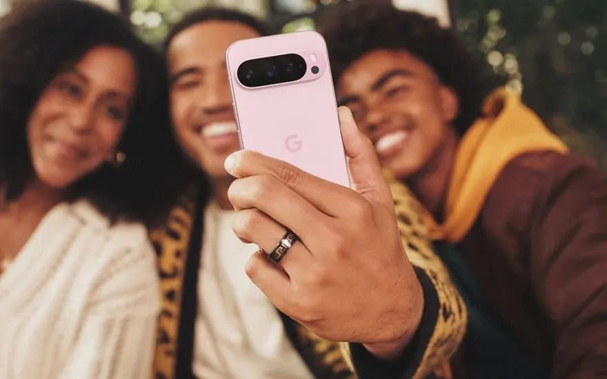 Three friends taking a selfie with a pink Google Pixel smartphone, all smiling happily in an outdoor environment.