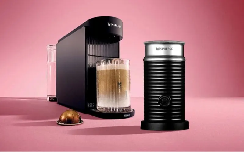 Zwarte Nespresso koffiemachine met een glas koffie en melkopschuimer op een roze achtergrond.