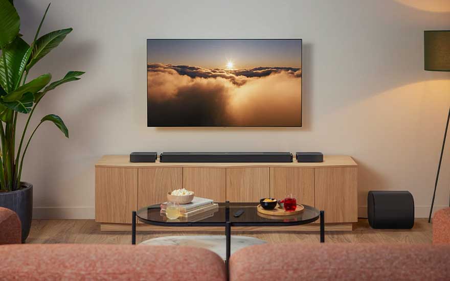 Rustige woonkamer met wand-tv, soundbar, twee kleine speakers, subwoofer, houten kast, planten en warm avondlicht.