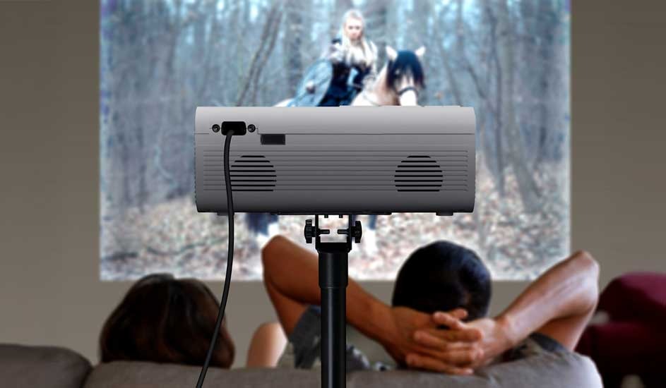 Two people are relaxing on a sofa, watching a film projected onto a wall. The projector is set up on a stand, but no brand name is visible.