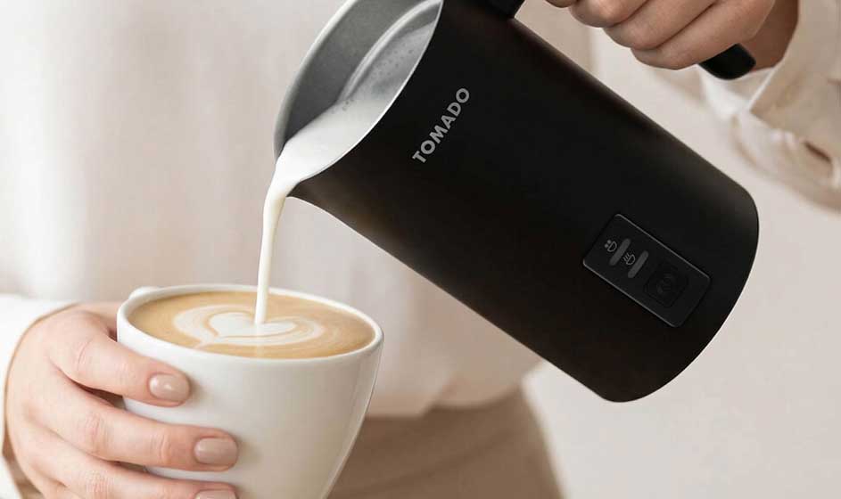 Een persoon schenkt melk uit een zwarte melkopschuimer in een kop koffie, terwijl een andere hand de witte kop stevig vasthoudt.