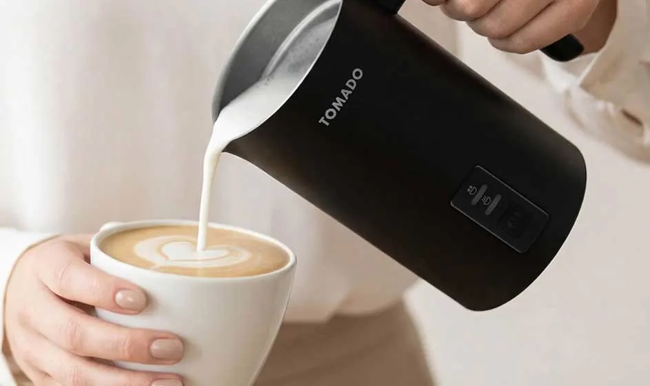 Een persoon schenkt melk uit een zwarte melkopschuimer in een kop koffie, terwijl een andere hand de witte kop stevig vasthoudt.