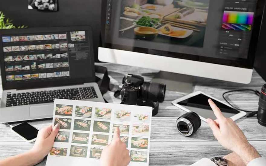Two people review printed food photos while editing images on a laptop and monitor, surrounded by a camera body and loose lenses.