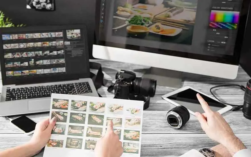 Two people review printed food photos while editing images on a laptop and monitor, surrounded by a camera body and loose lenses.