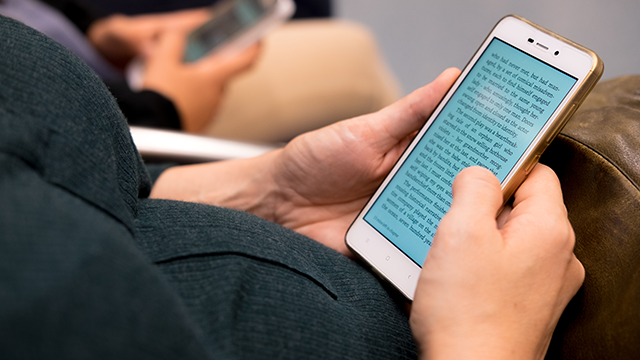 Een persoon houdt een witte smartphone met een blauw scherm vast, waarop tekst staat, terwijl ze op een bruine leren stoel zitten.