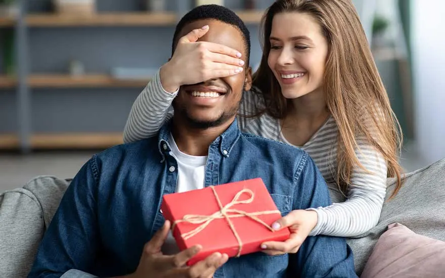 Een man zit op de bank met een rood geschenk; zijn partner bedekt lachend zijn ogen van achteren om hem een lieve verrassing te geven.