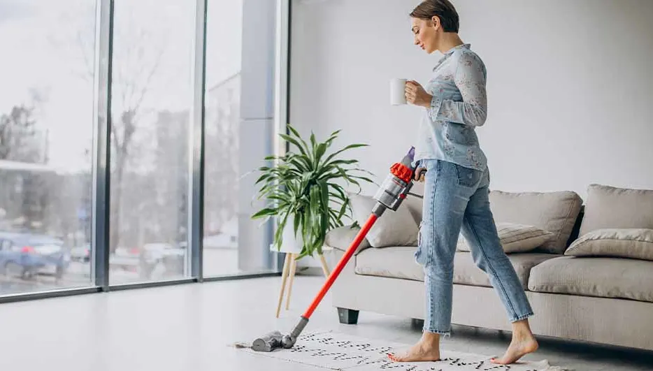 Vrouw stofzuigt een kleed in een lichte kamer met een grote plant en een beige bank, kopje in hand. Grote ramen op achtergrond.