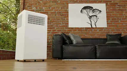 Een witte airconditioner staat in een kamer met een bakstenen muur, een zwarte bank en een schilderij van bomen.