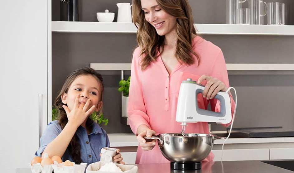 Een vrouw mixt in een kom met een handmixer; naast haar likt een meisje deeg van haar vingers, met eieren en bakspullen op het aanrecht.