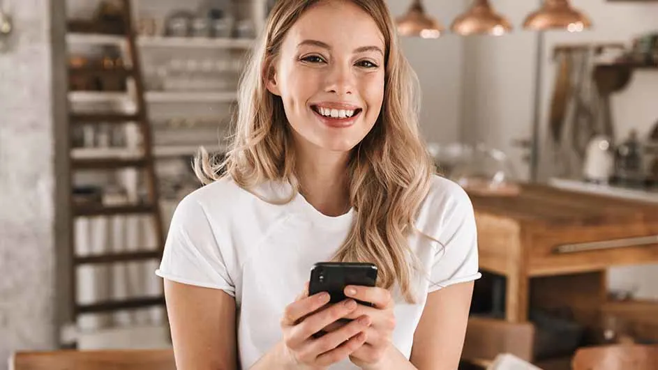 Jonge vrouw glimlacht, houdt een zwarte smartphone vast. Achtergrond: keuken met houten elementen, witte kasten en koperen hanglampen.