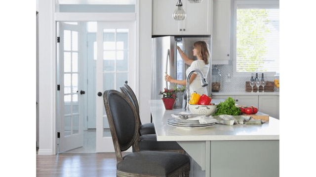 Een keuken met een vrouw die de koelkast opent. Op het aanrecht staan groenten en borden. Er staan twee stoelen aan het aanrecht.
