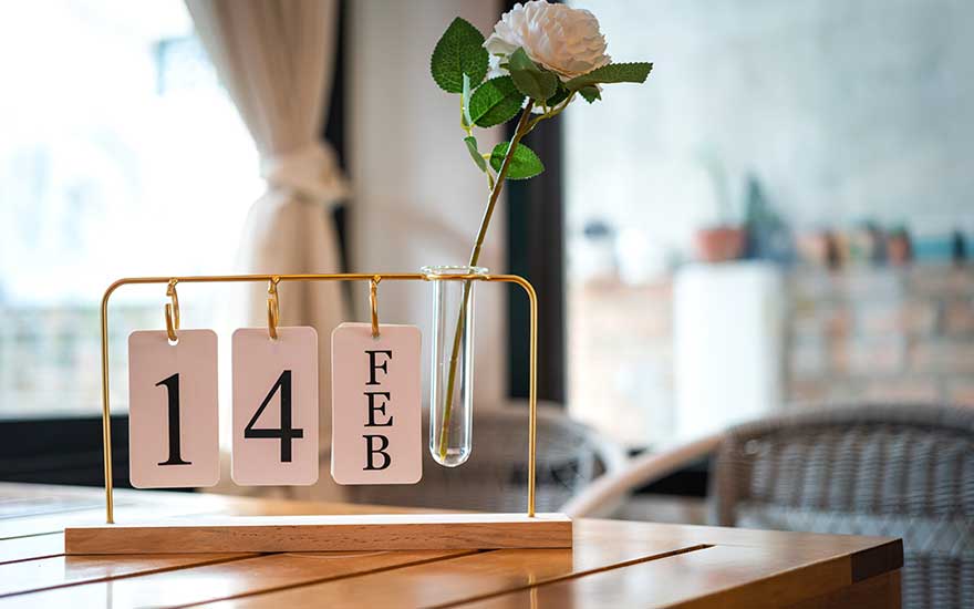 Op een houten tafel staat een stijlvol kalenderrek met de datum 14 FEB naast een glazen buisvaasje met een enkele witte roos.  EN: On a wooden table, a stylish calendar shows 14 FEB beside a slim glass tube vase holding a single white rose in soft daylight.  FR: Sur une table en bois, un élégant calendrier affiche le 14 FÉV, posé à côté d’un fin vase en verre contenant une rose blanche.