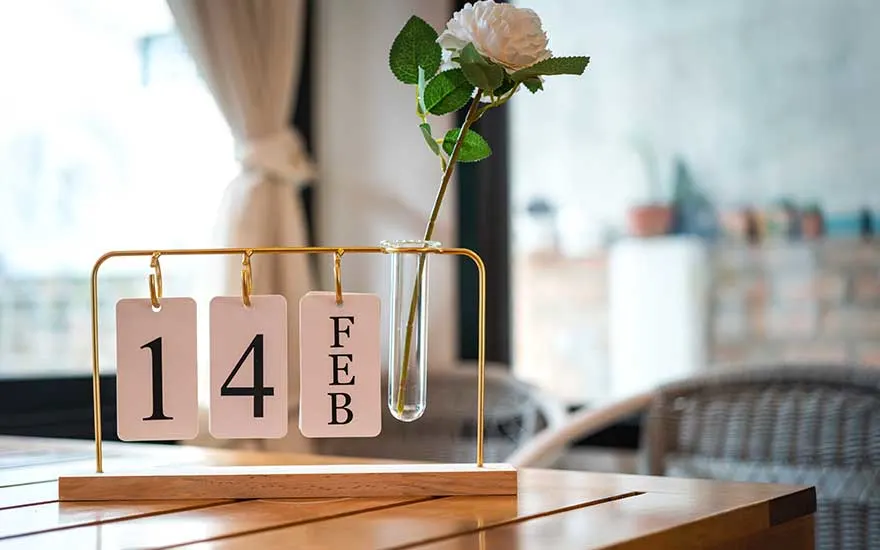 Op een houten tafel staat een stijlvol kalenderrek met de datum 14 FEB naast een glazen buisvaasje met een enkele witte roos. EN: On a wooden table, a stylish calendar shows 14 FEB beside a slim glass tube vase holding a single white rose in soft daylight. FR: Sur une table en bois, un élégant calendrier affiche le 14 FÉV, posé à côté d’un fin vase en verre contenant une rose blanche.