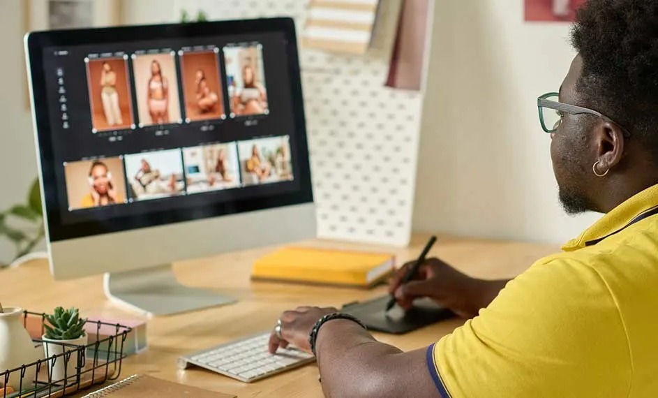 Designer editing fashion photos on a large desktop screen, using a stylus and keyboard on a neatly arranged wooden desk.