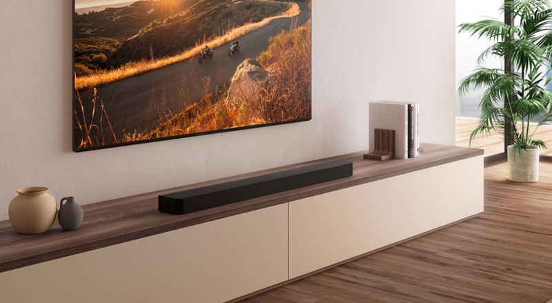 Rustige hoek van de kamer met lang houten sideboard en witte laden, daarop een zwarte soundbar en stapel boeken, erboven een wand-gemonteerde tv met wegdekscène, naast het meubel een grote potplant.