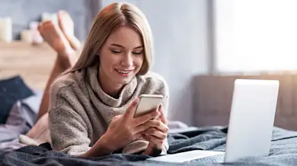 Vrouw ligt op een bed met een telefoon in haar handen, naast een witte laptop, op een grijze deken.