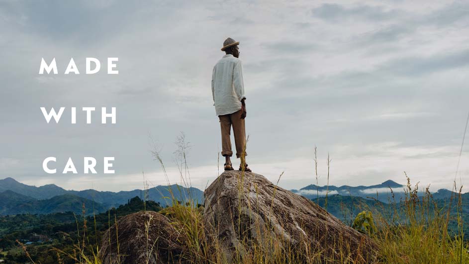 Man staat op een rots met een hoed, een wit hemd en een bruine broek, kijkend naar heuvels onder een bewolkte lucht. Tekst: "Made With Care."