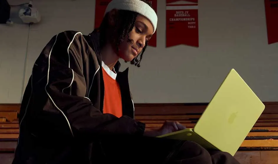 Student sits on wooden bleachers in a sports hall, wearing a track jacket and beanie, typing intently on an open yellow laptop on their lap.