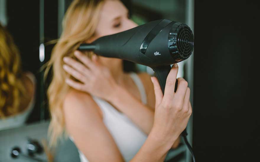 Vrouw met golvend blond haar gebruikt een matzwarte föhn in de badkamer; ze richt de luchtstroom op haar haar terwijl ze het met de hand ordent.