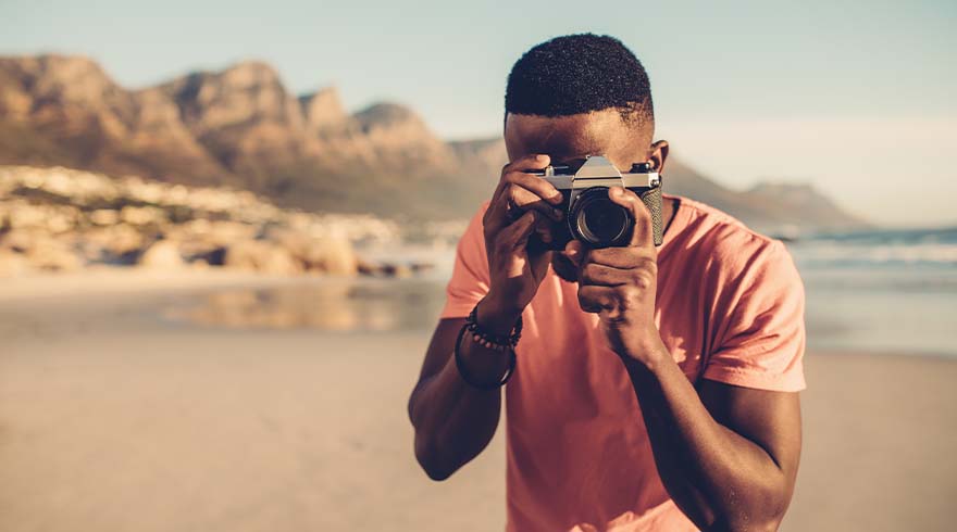 Een persoon fotografeert met een camera op het strand, met bergen en de zee op de achtergrond.