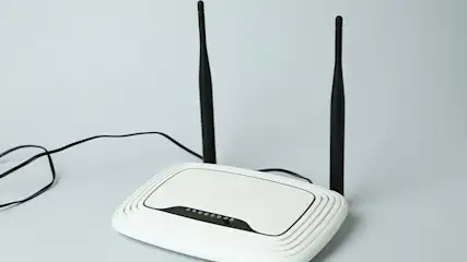 Een witte router met twee zwarte antennes staat op een lichtgrijze achtergrond. Een zwarte kabel is aangesloten op de router.