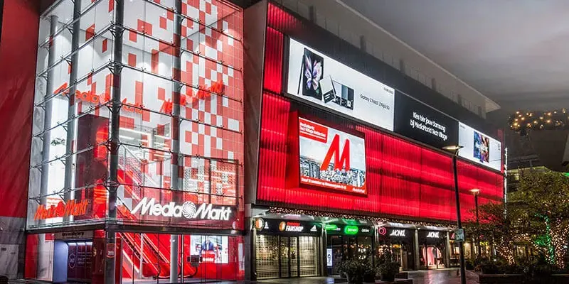 Een groot Media Markt gebouw met rode en witte reclamepanelen en diverse winkels op de begane grond.