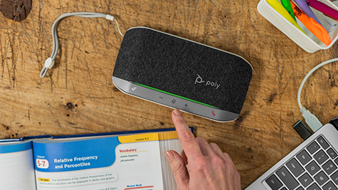 Hand bedient een Poly speakerphone op houten bureau, naast open studieboek, laptop en kleurrijke markeerstiften voor online leren.