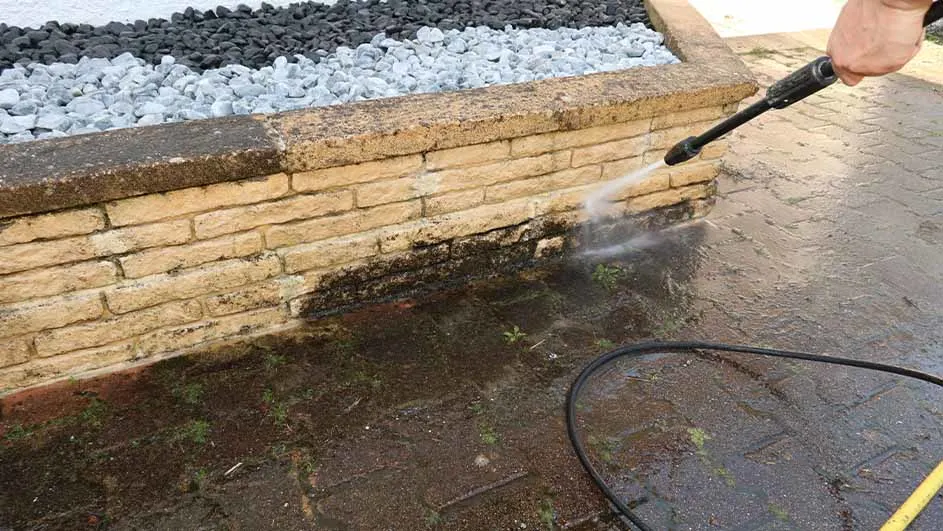Persoon gebruikt een hogedrukreiniger om vuil en algen van een gemetselde tuinmuur te verwijderen, naast decoratieve witte en zwarte stenen.