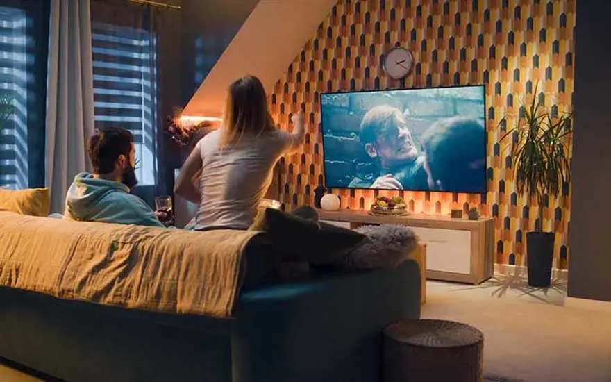 Two people sit together on a sofa, watching an intense film or series on a large TV in a stylish and warmly lit room.