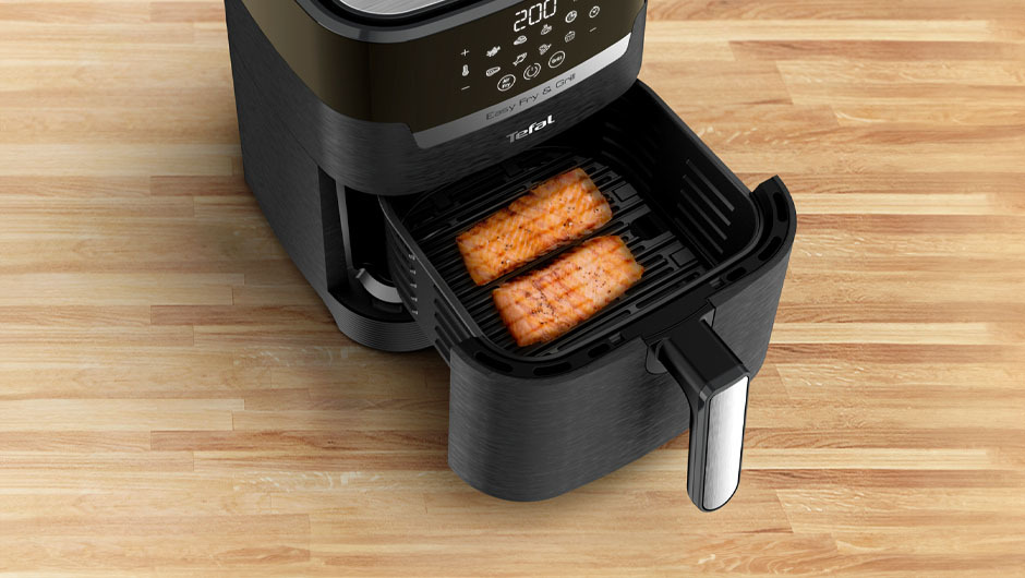 Een zwarte airfryer met een lade open, waarin twee stukken vis op een rooster liggen, op een houten oppervlak.